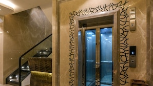 A modern elevator with a glass door, a marble wall with a geometric pattern, and a staircase with a black railing. - Hotel Orient Regency (Mumbai)