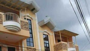 Edificio residencial de varios pisos con balcones, entrada protegida por un portón y un cartel que dice 'House for Sale'.