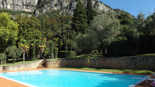 A swimming pool with stone steps leading down to the water, surrounded by a stone wall and lush greenery. - Villa delle Sirene (Garda)