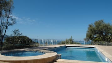 A swimming pool with a hot tub, surrounded by a fence and trees, with a clear sky in the background.