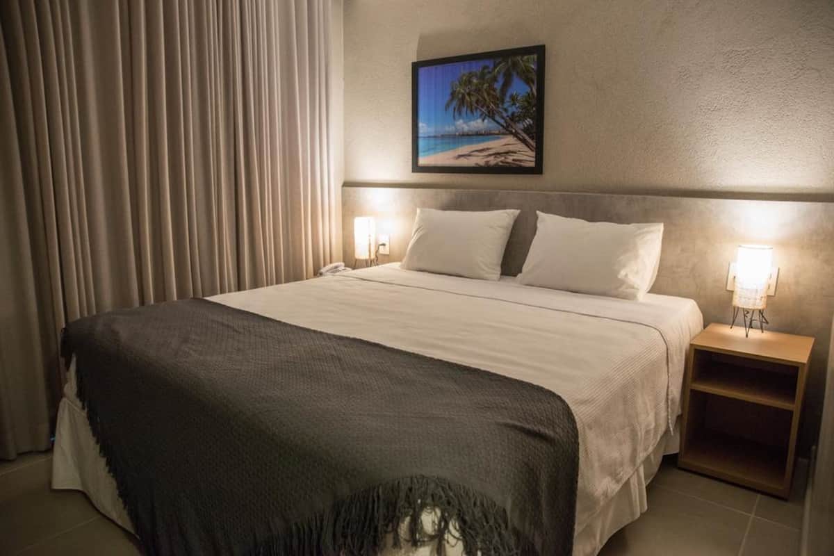 A hotel room with a bed, a nightstand with a lamp, a framed picture on the wall, and a window with curtains.