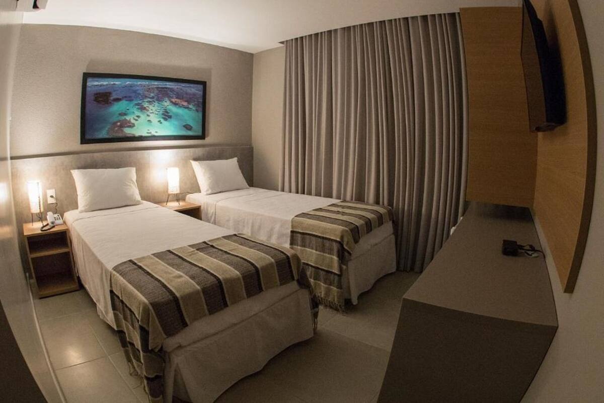 A hotel room with two beds, a TV mounted on the wall, a desk, and a large window with curtains.