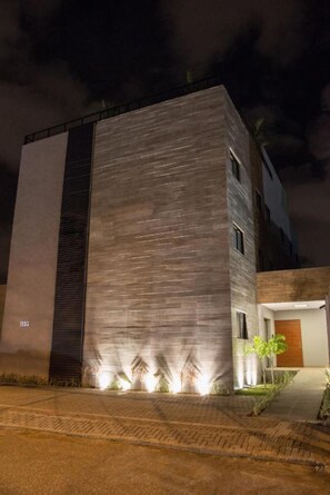 Un edificio moderno con techo plano, una sección vertical grande y una entrada pequeña con puerta y un árbol pequeño.