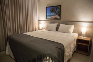 A hotel room with a bed, a nightstand with a lamp, a framed picture on the wall, and a window with curtains.