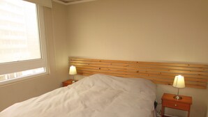 A bedroom with a wooden headboard, two bedside tables with lamps, a bed with white bedding, and a window with blinds. - 9.- HNA Mirador El Faro (La Serena)