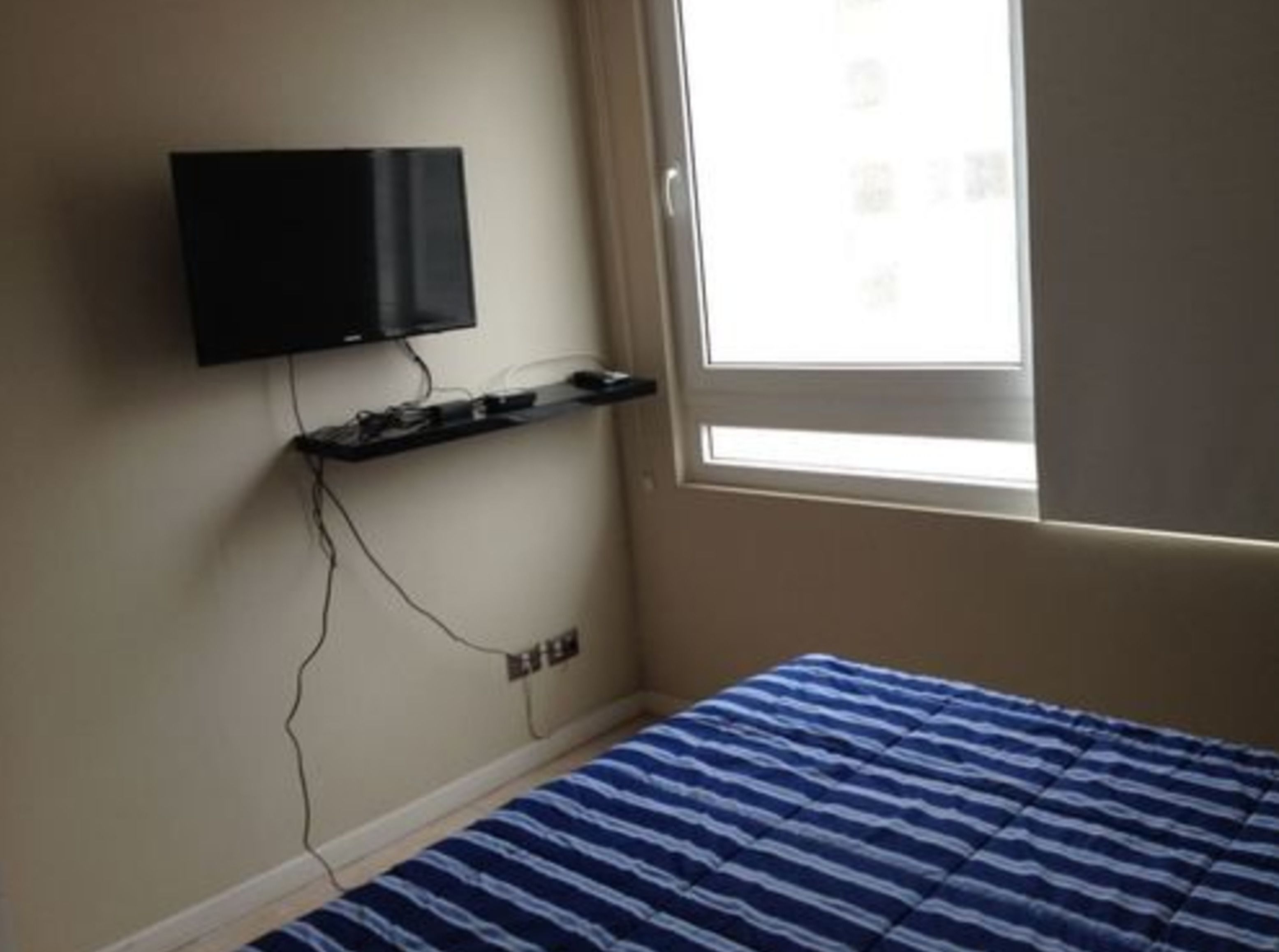 Une chambre avec un lit, un téléviseur fixé au mur et une fenêtre avec des stores.