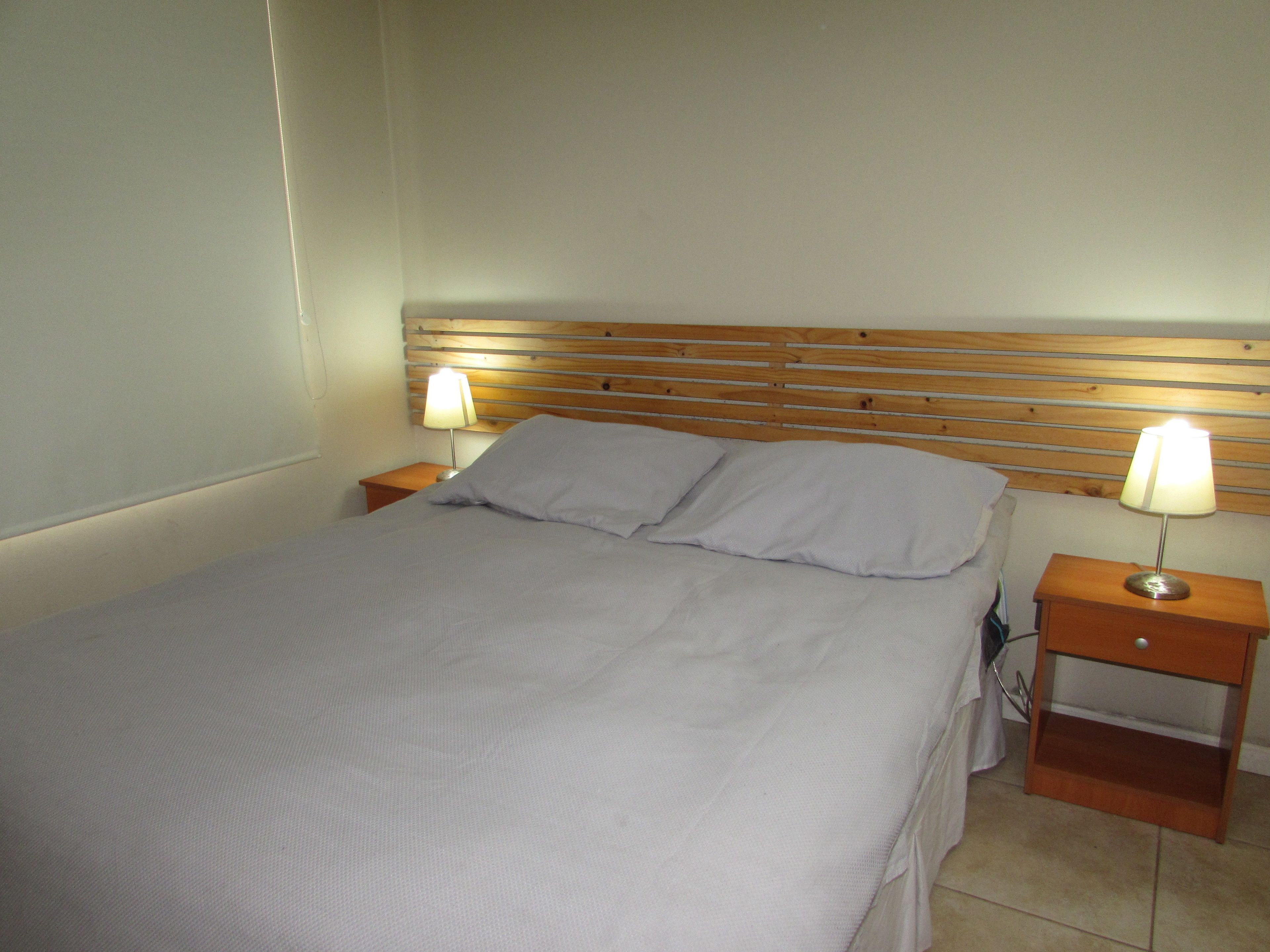 A neatly made bed with a wooden headboard, two bedside tables with lamps, and a plain wall.