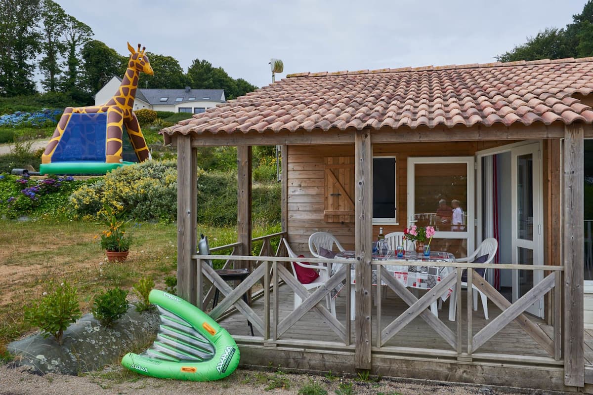 Een houten chalet met pannendak, een overdekt terras met tafel en stoelen, en een glijbaan op de achtergrond.