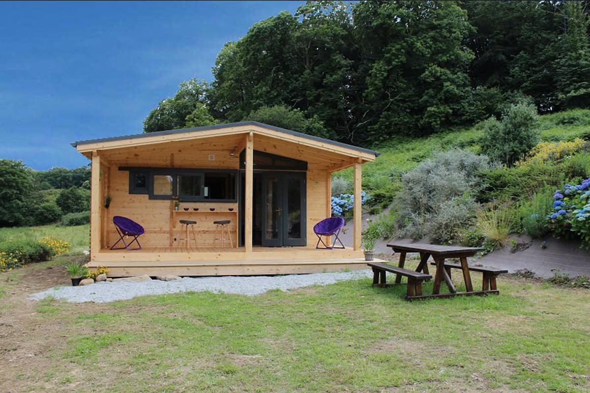 Een houten chalet met een klein terras, twee barkrukken, een paarse stoel en een picknicktafel.