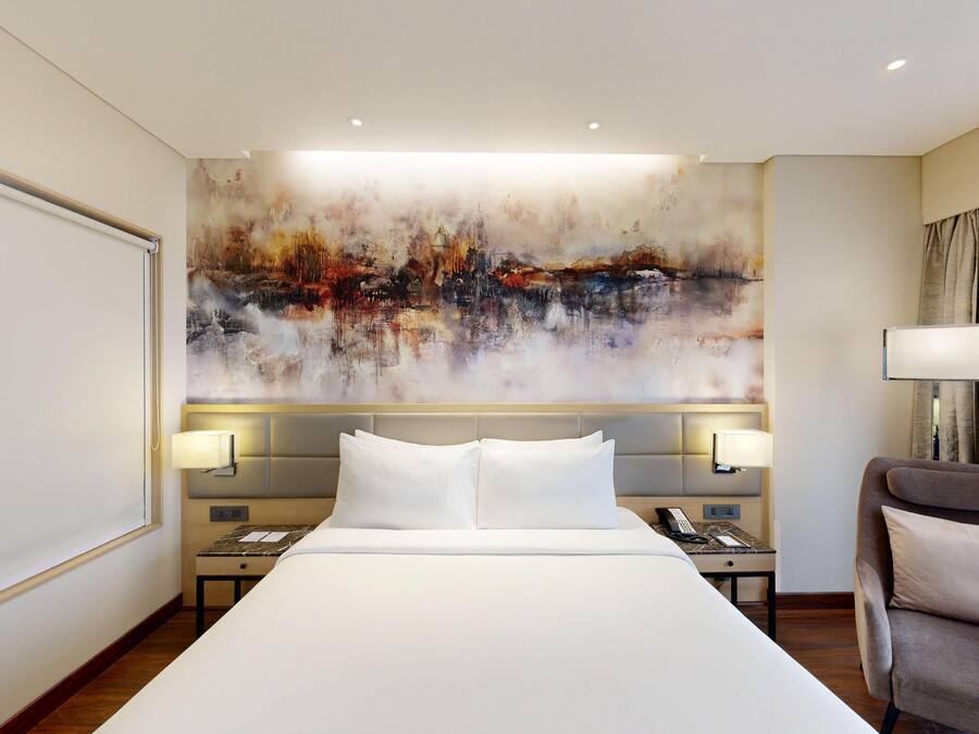 Un cuarto de hotel con una cama grande, dos mesitas de noche con lámparas, una silla y un cuadro abstracto de gran tamaño en la pared.