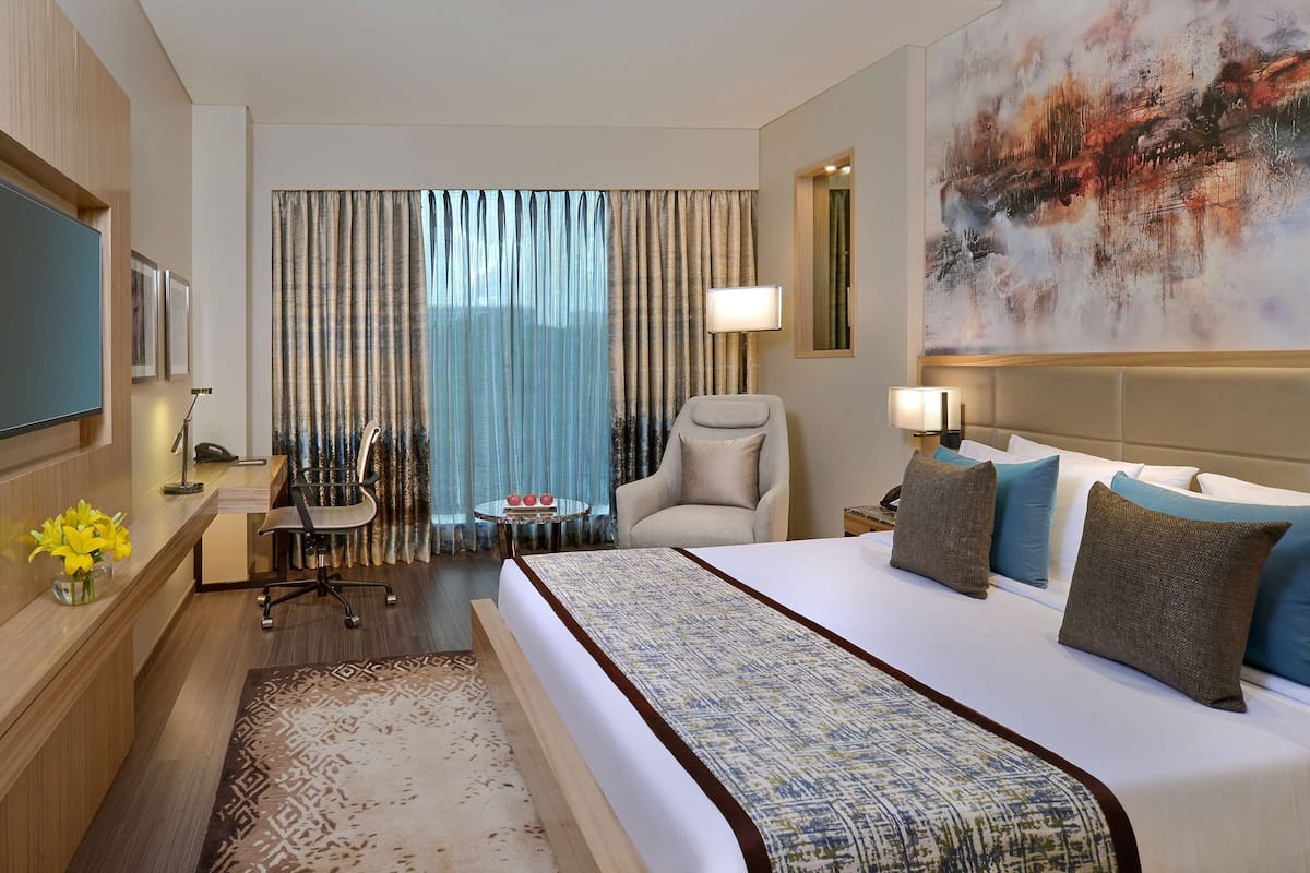 A hotel room with a bed, a desk with a chair, a TV, a lamp, and a painting on the wall.
