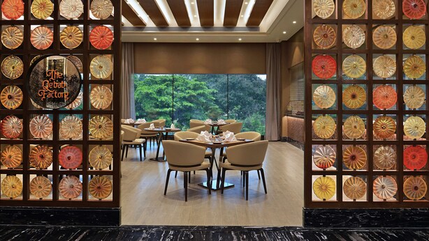 A restaurant with a glass wall displaying numerous decorative plates, a sign reading "The Deba Factory," and a view of trees through large windows. - Radisson Mumbai Andheri MIDC (Mumbai)
