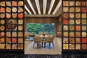 A restaurant with a glass wall displaying numerous decorative plates, a sign reading "The Deba Factory," and a view of trees through large windows. - Radisson Mumbai Andheri MIDC (Mumbai)
