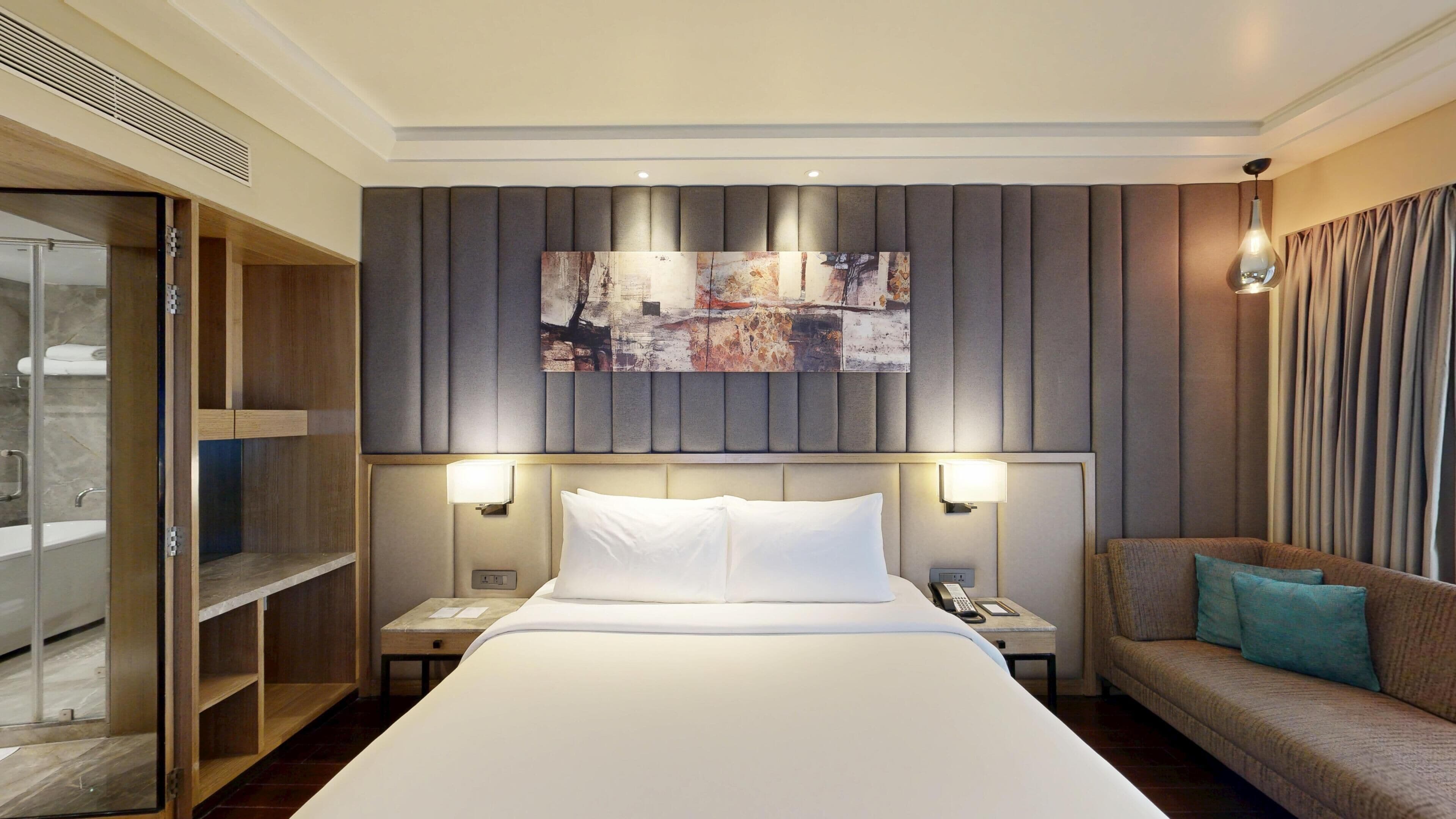 A hotel room with a large bed, a sofa, a desk with a lamp, a painting on the wall, and a walk-in closet.