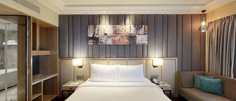 A hotel room with a large bed, a sofa, a desk with a lamp, a painting on the wall, and a walk-in closet.