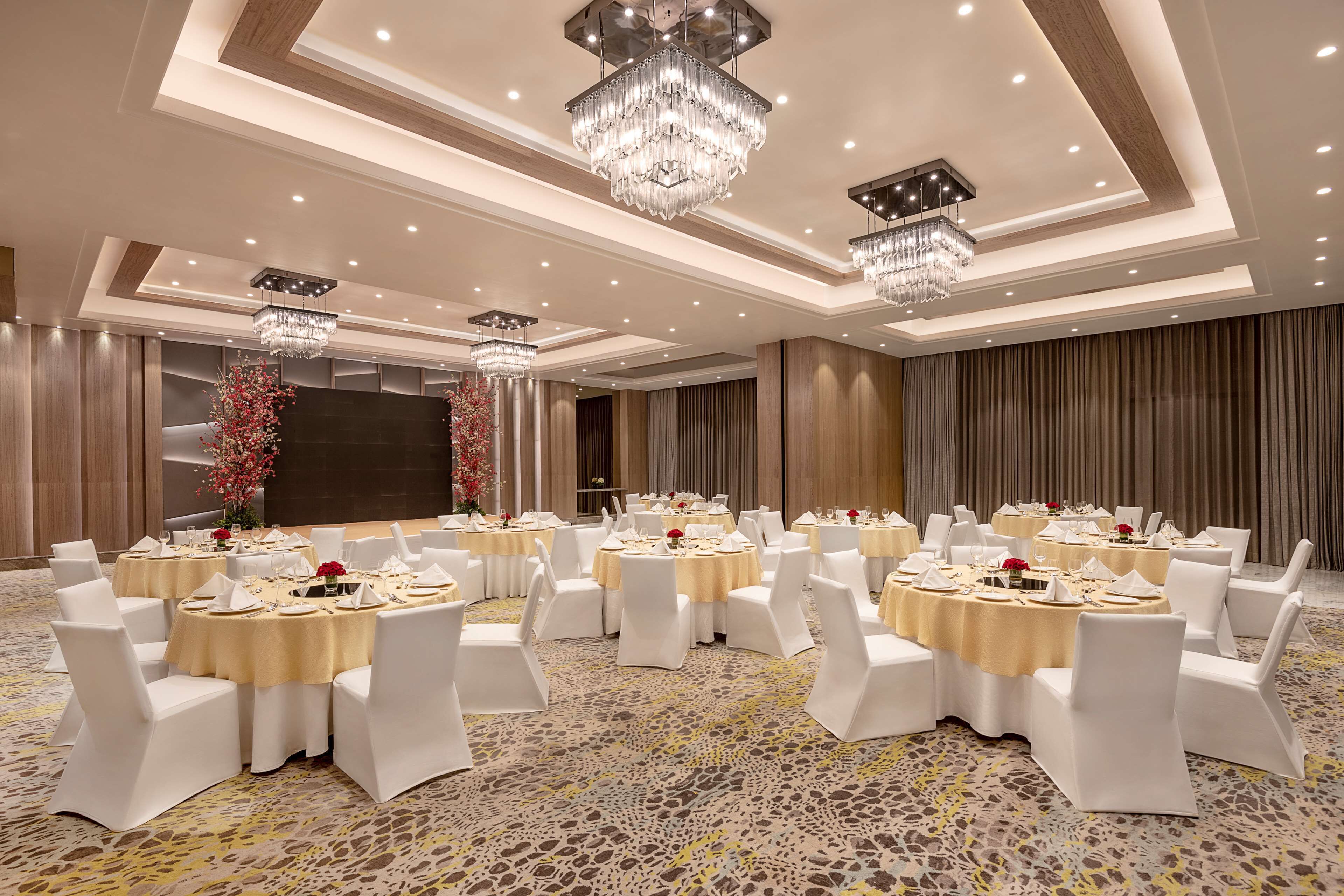 A banquet hall with round tables, white chairs, chandeliers, and a patterned carpet.