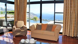 Ein Wohnzimmer mit einem beigen Sofa, einem Sessel und einem Couchtisch mit Dekoration. Große Fenster bieten Blick auf das Meer und eine weite Landschaft.