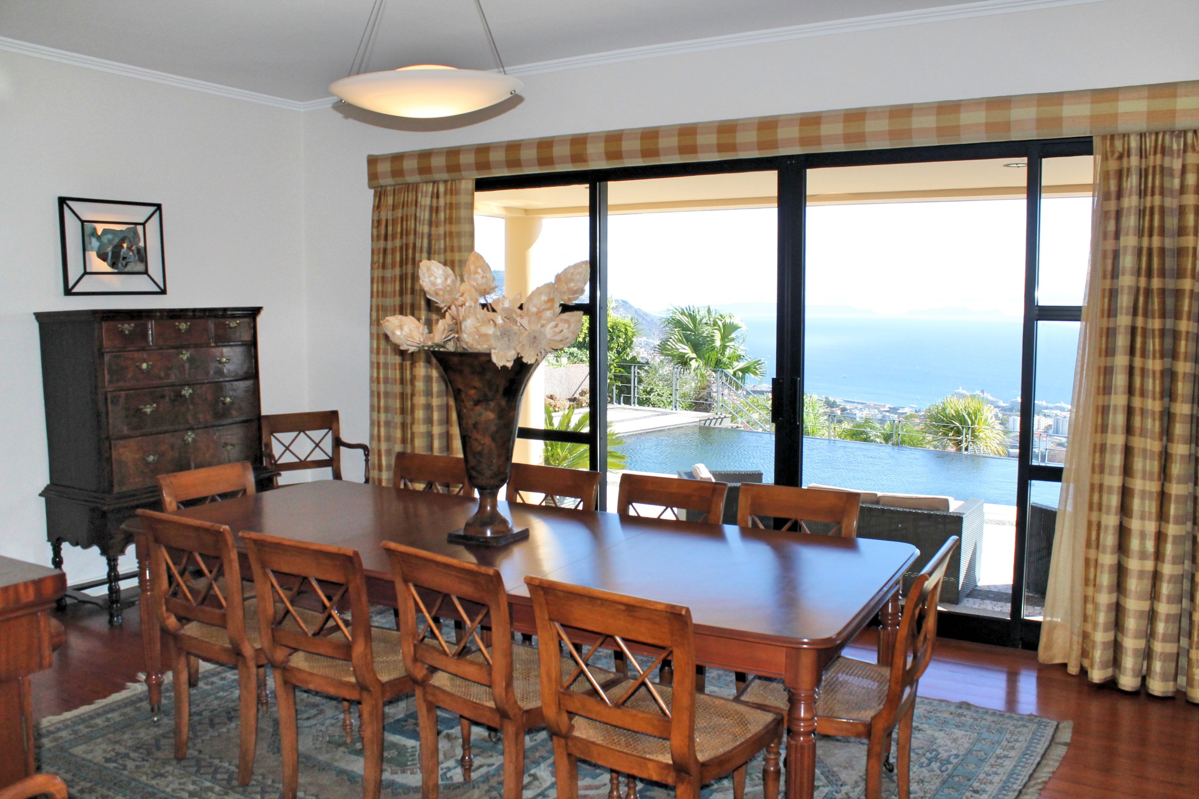 A dining area with a table and chairs, a vase with flowers, a wooden cabinet, and a view of the ocean through large windows.