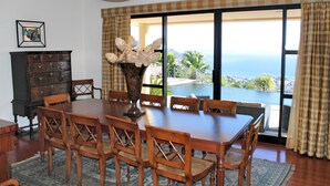 Ein Essbereich mit Tisch und Stühlen, einer Vase mit Blumen, einer Holzkommode und Blick auf das Meer durch große Fenster.