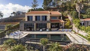 Ein modernes, zweistöckiges Haus mit Swimmingpool, Sitzgelegenheiten im Freien und Blick auf eine Hügellandschaft.