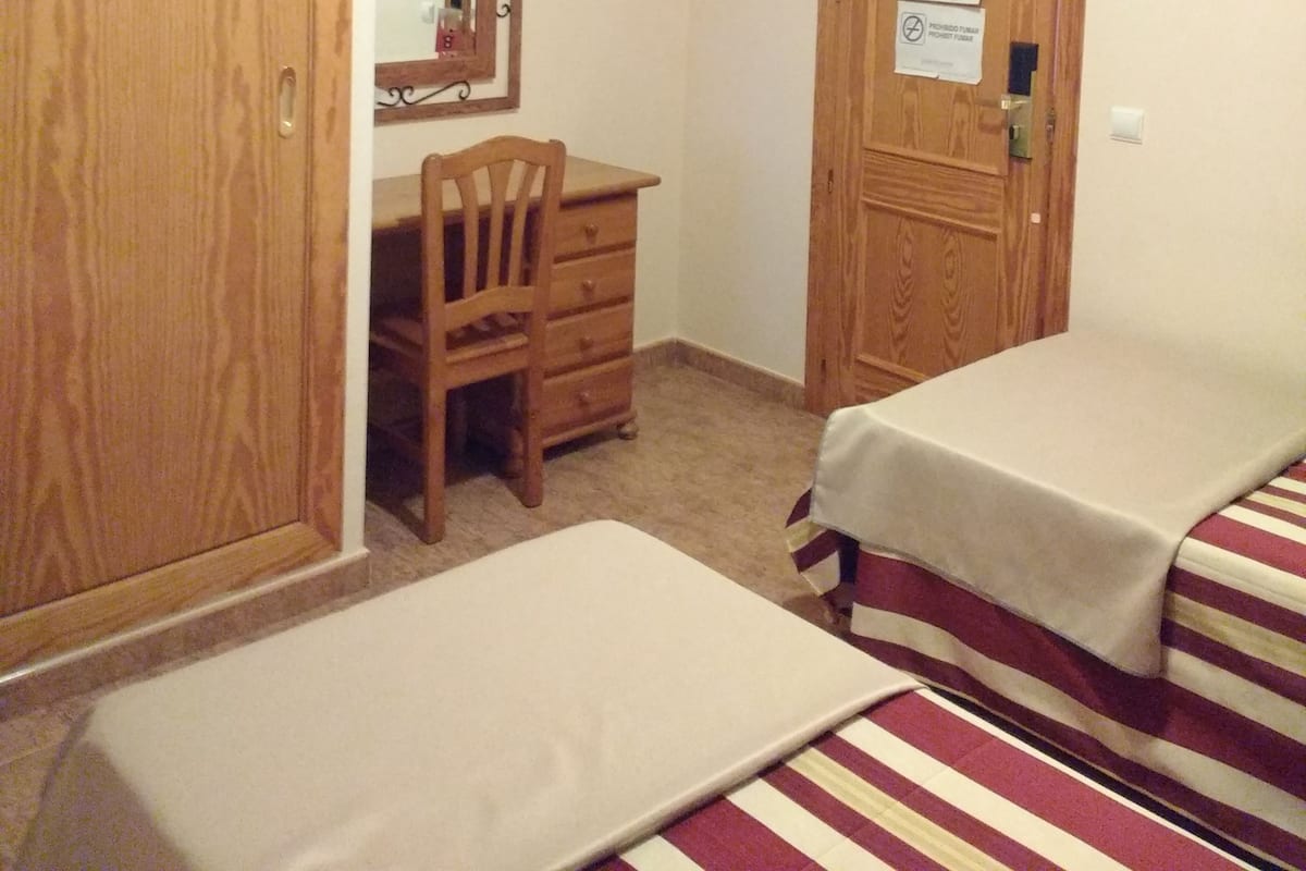 Een kamer met een houten garderobe, een bureau met stoel, een aan de muur bevestigde televisie en twee bedden met gestreepte beddengoed.