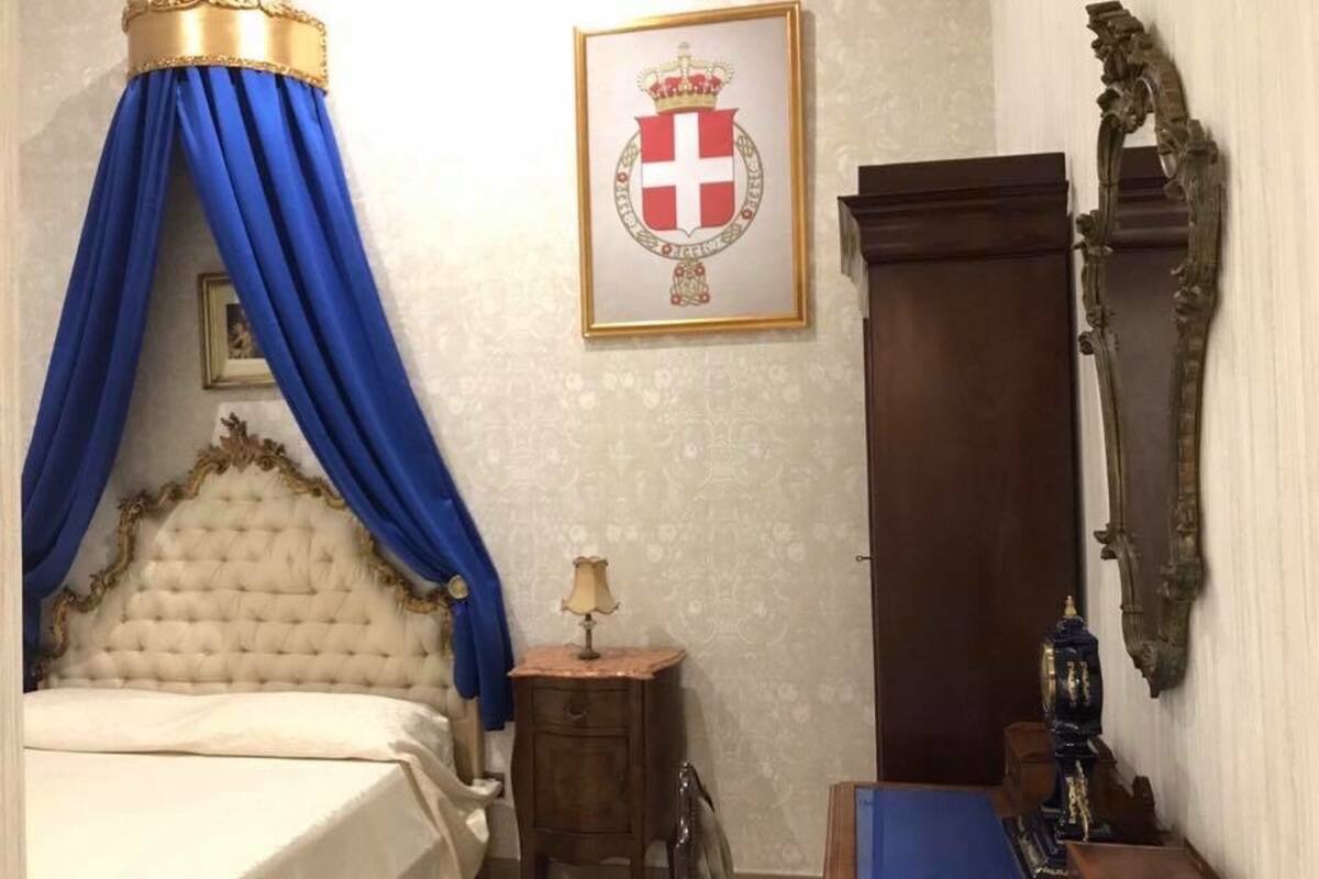 A bedroom with a four-poster bed, a framed coat of arms on the wall, a wooden wardrobe, and a small bedside table with a lamp.