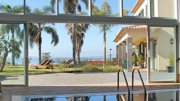 A pool area with clear views of palm trees and a beach, and a building with a tiled roof and columns. - Villa Luzia by Our Madeira (Funchal)