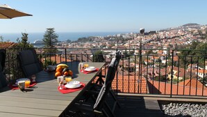 A balcony with a table set for breakfast, overlooking a cityscape with red-roofed buildings and a view of the sea. - Villa Luzia by Our Madeira (Funchal)