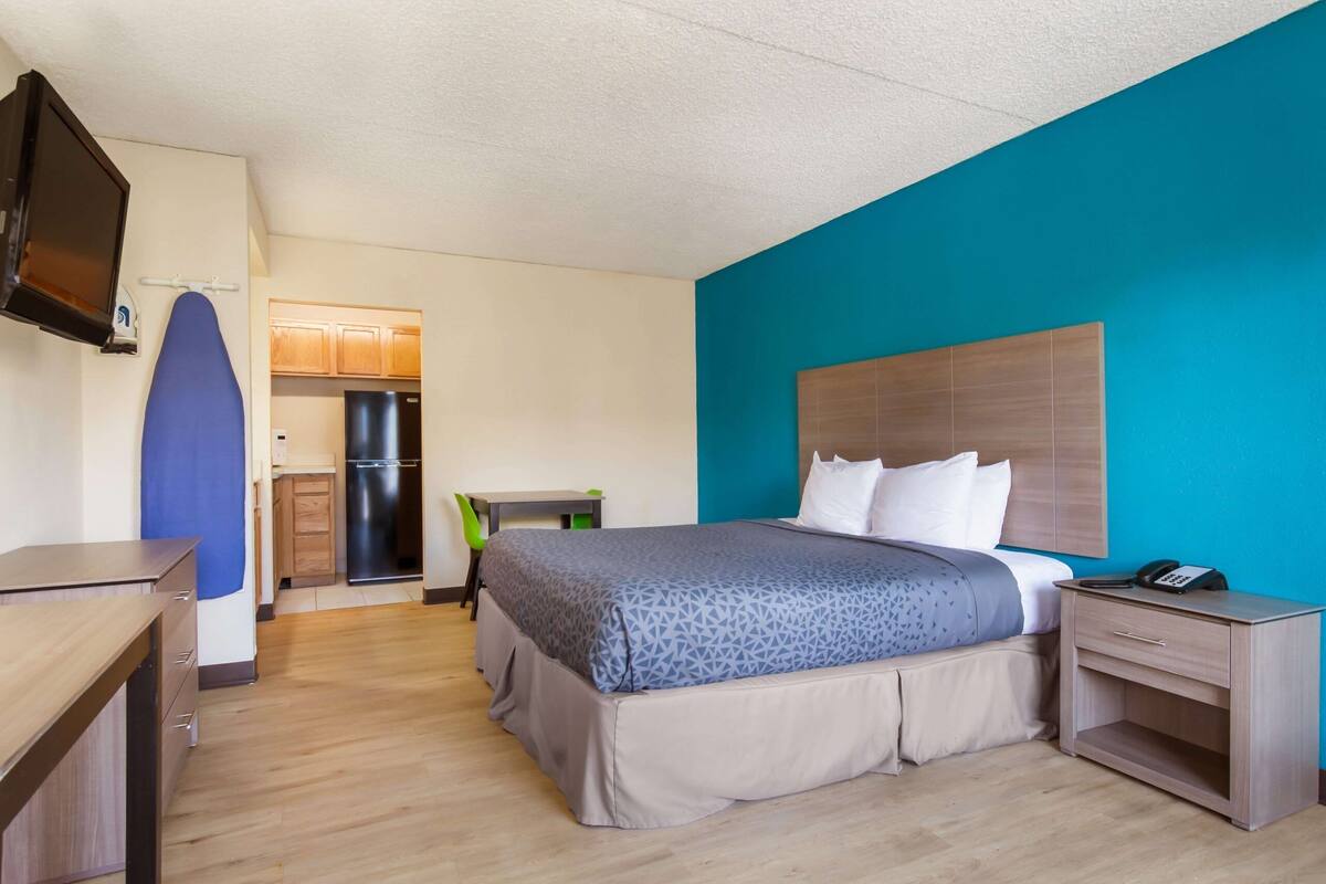 A hotel room with a bed, a desk, a chair, a TV, and a kitchenette.