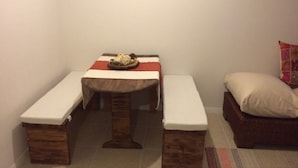 Een kleine, gezellige kamer met een houten tafel en bankjes, een bed met kussen, en een vloerkleed.