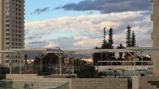 Een terras op het dak met een glazen balustrade, een stenen muur en uitzicht op een stadsbeeld met een hoog gebouw en bomen.