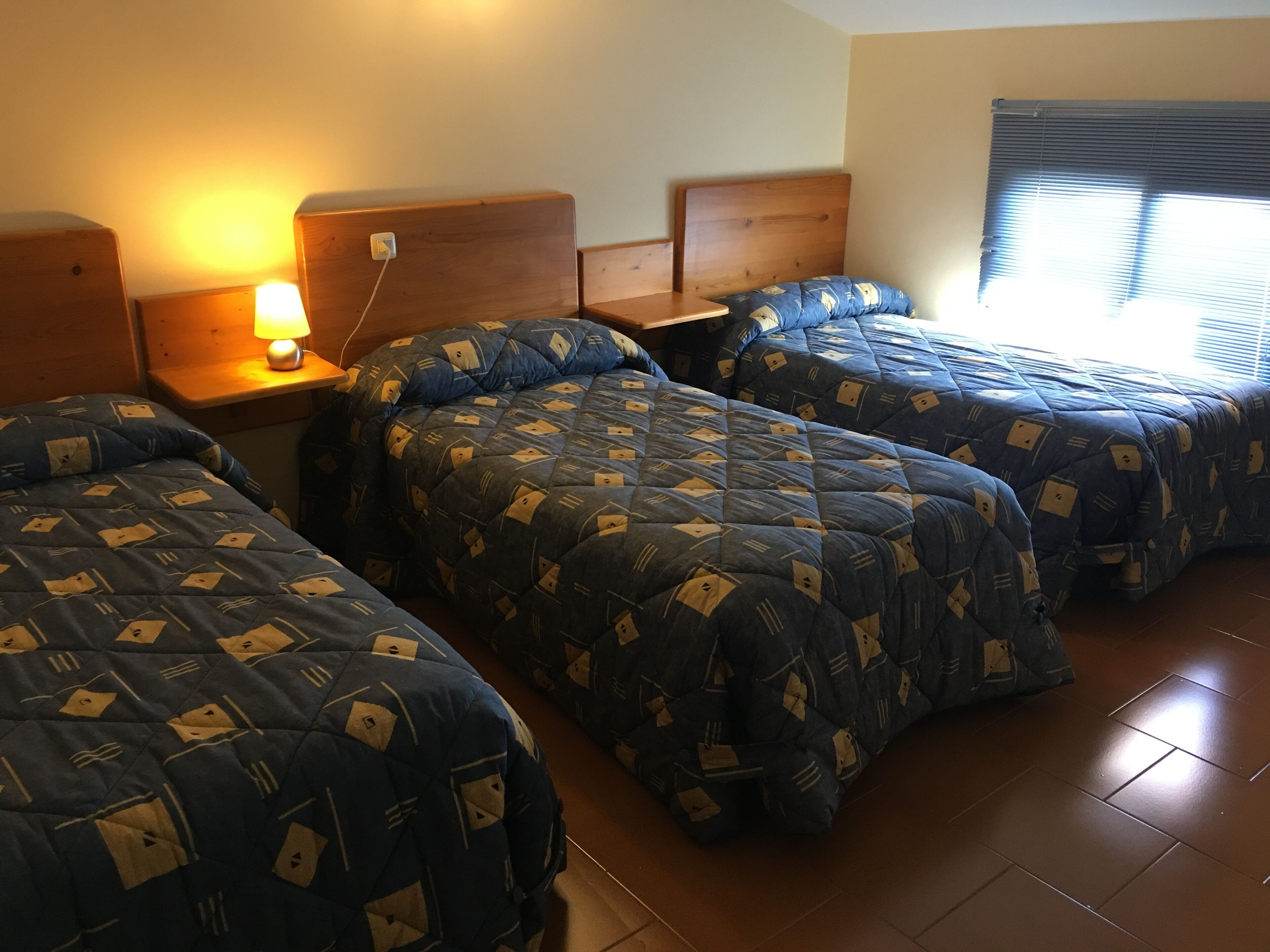 A room with three beds, each with a blue quilted cover featuring a geometric pattern. There is a bedside table with a lamp on the left side, and a window with blinds on the right.