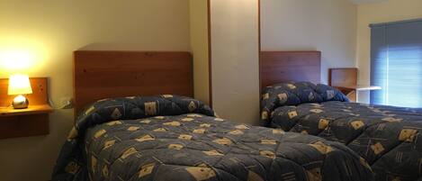 A bedroom with two beds, each with a wooden headboard, a bedside table with a lamp, and a window with blinds.
