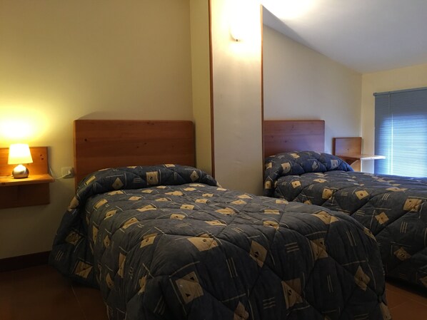 A bedroom with two beds, each with a wooden headboard, a bedside table with a lamp, and a window with blinds.