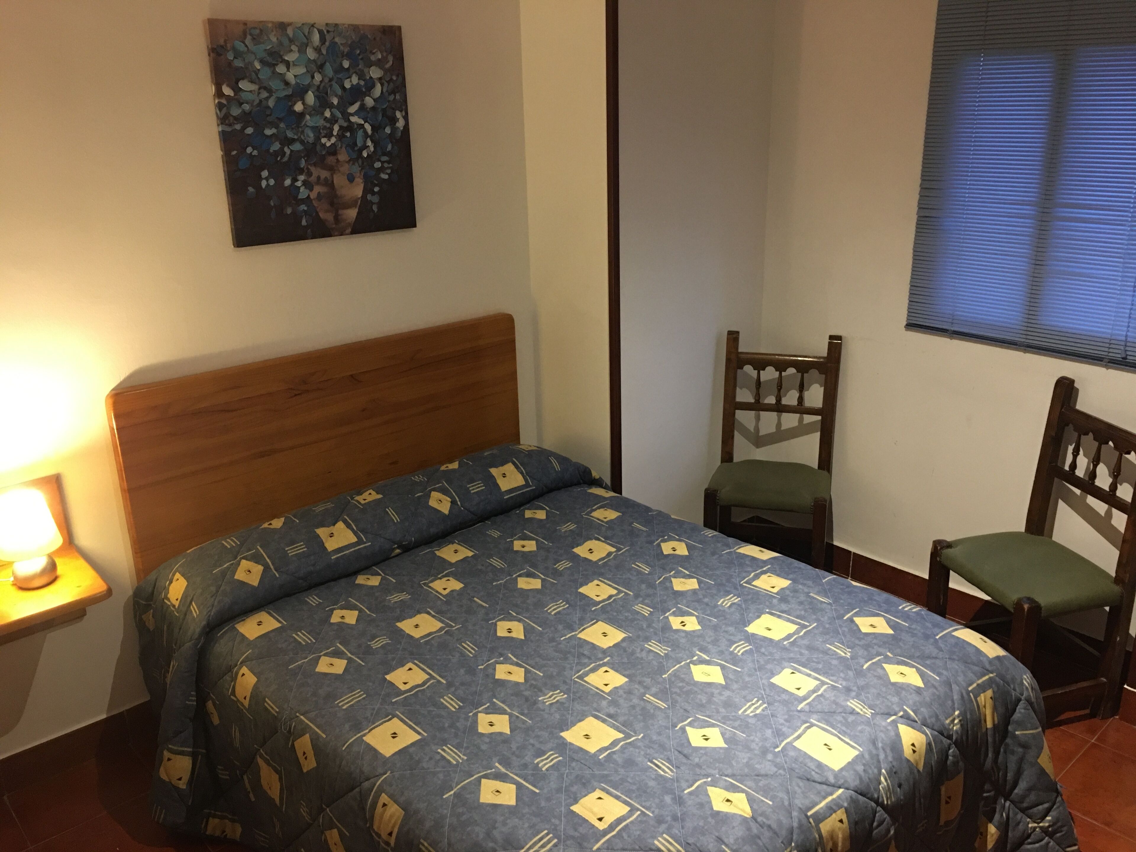 A bedroom with a wooden headboard, a bed with a patterned blue and yellow bedspread, a small table with a lamp, a chair, and a painting on the wall.