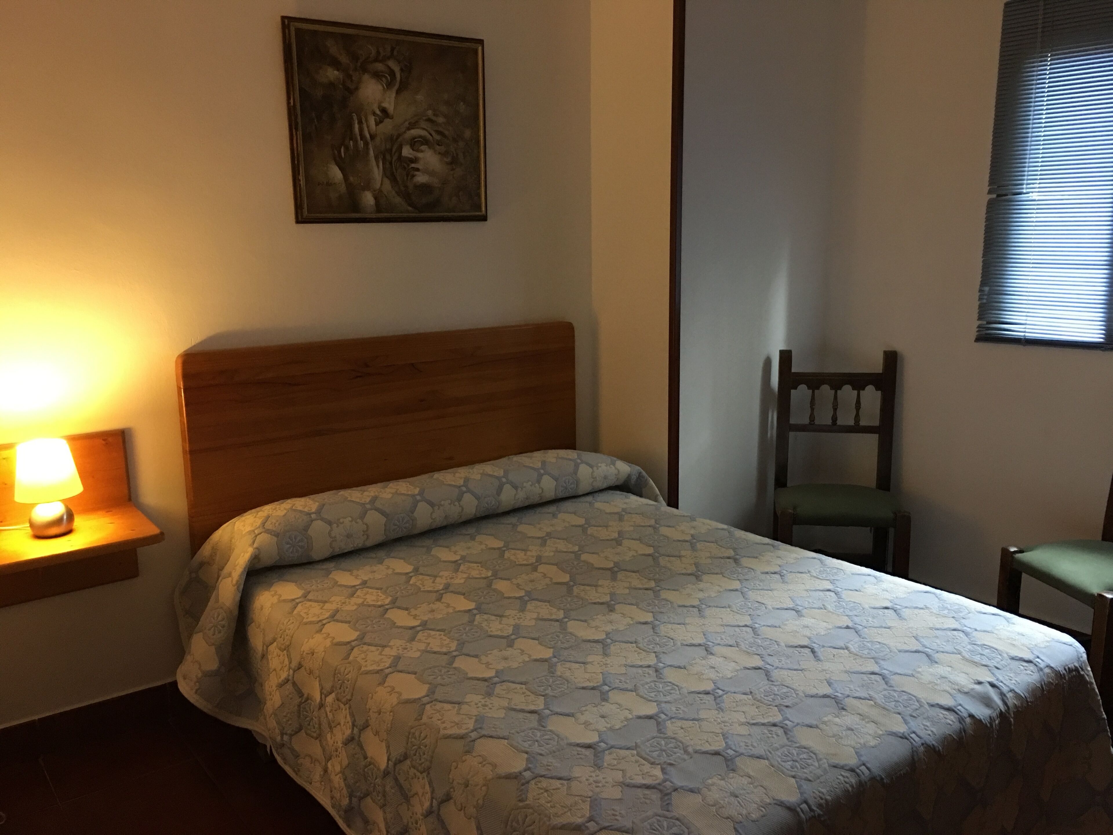 A bedroom with a wooden headboard, a patterned bedspread, a nightstand with a lamp, a chair, and a painting on the wall.