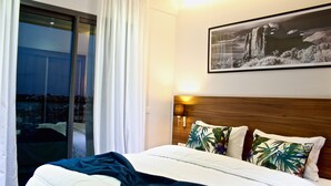Habitación de hotel con cama, mesita de noche con lámpara, ventana con cortinas e imagen enmarcada en la pared.