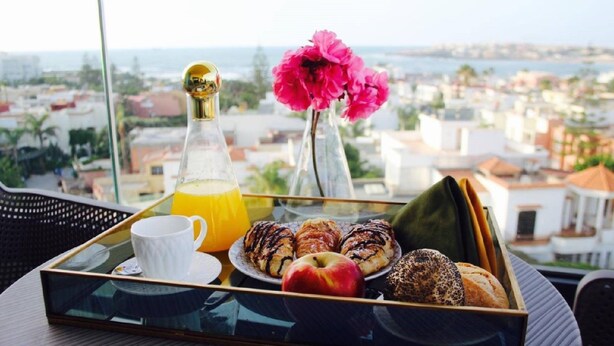 Bandeja de desayuno con pastelería, una manzana y jugo de naranja en un balcón con vista a la ciudad.