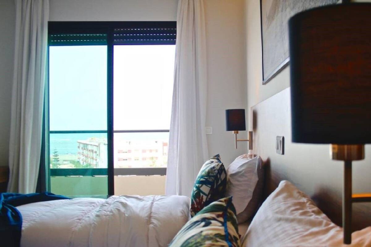 A hotel room with a bed, a nightstand, a lamp, and a view of the sea through a window.
