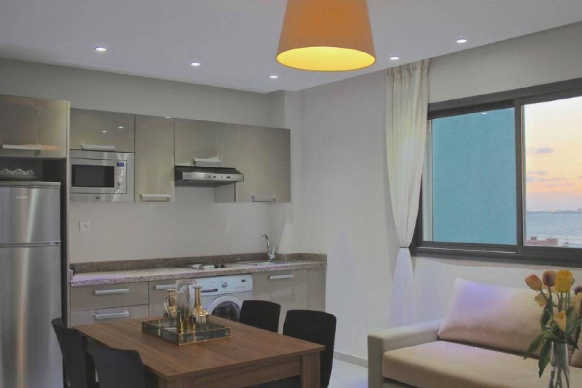 A modern kitchen with stainless steel appliances, a dining area with a wooden table, and a sofa by a window with a view of the sea.