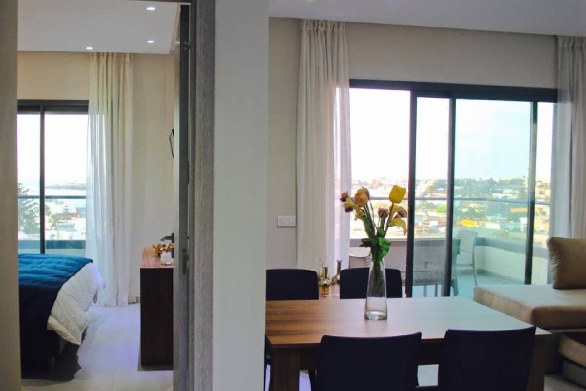 A hotel room with a bed, a dining table with chairs, a vase with flowers, and a balcony with a view.