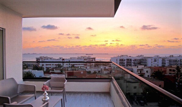 A balcony with a view of a sunset over the sea, a table with chairs, and a vase of flowers.