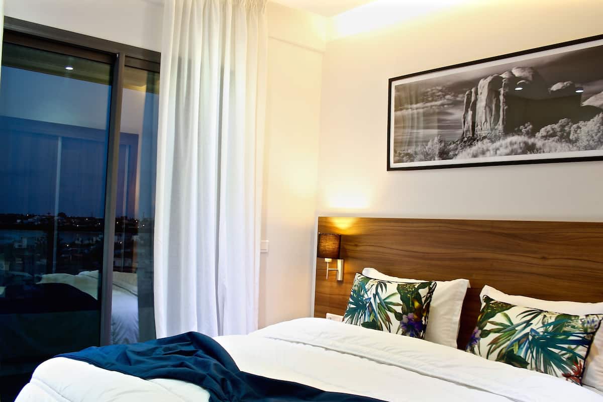 A hotel room with a bed, a nightstand, a wall-mounted lamp, a large window with curtains, and a framed picture on the wall.