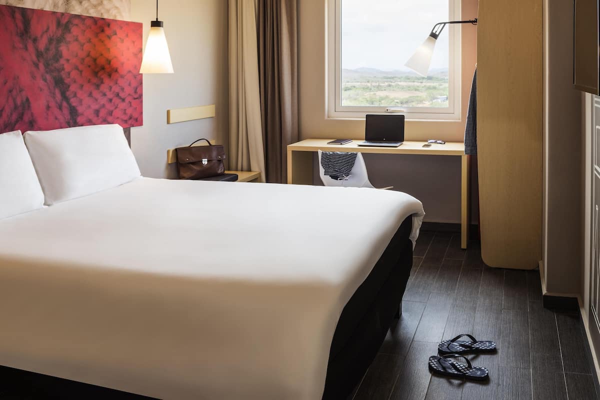 A hotel room with a bed, a desk with a laptop, a chair, a window with a view of the countryside, and a handbag on the nightstand.