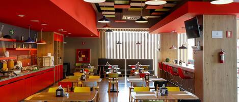 A modern restaurant interior with red walls, colorful ceiling panels, and a variety of seating arrangements including tables and booths.