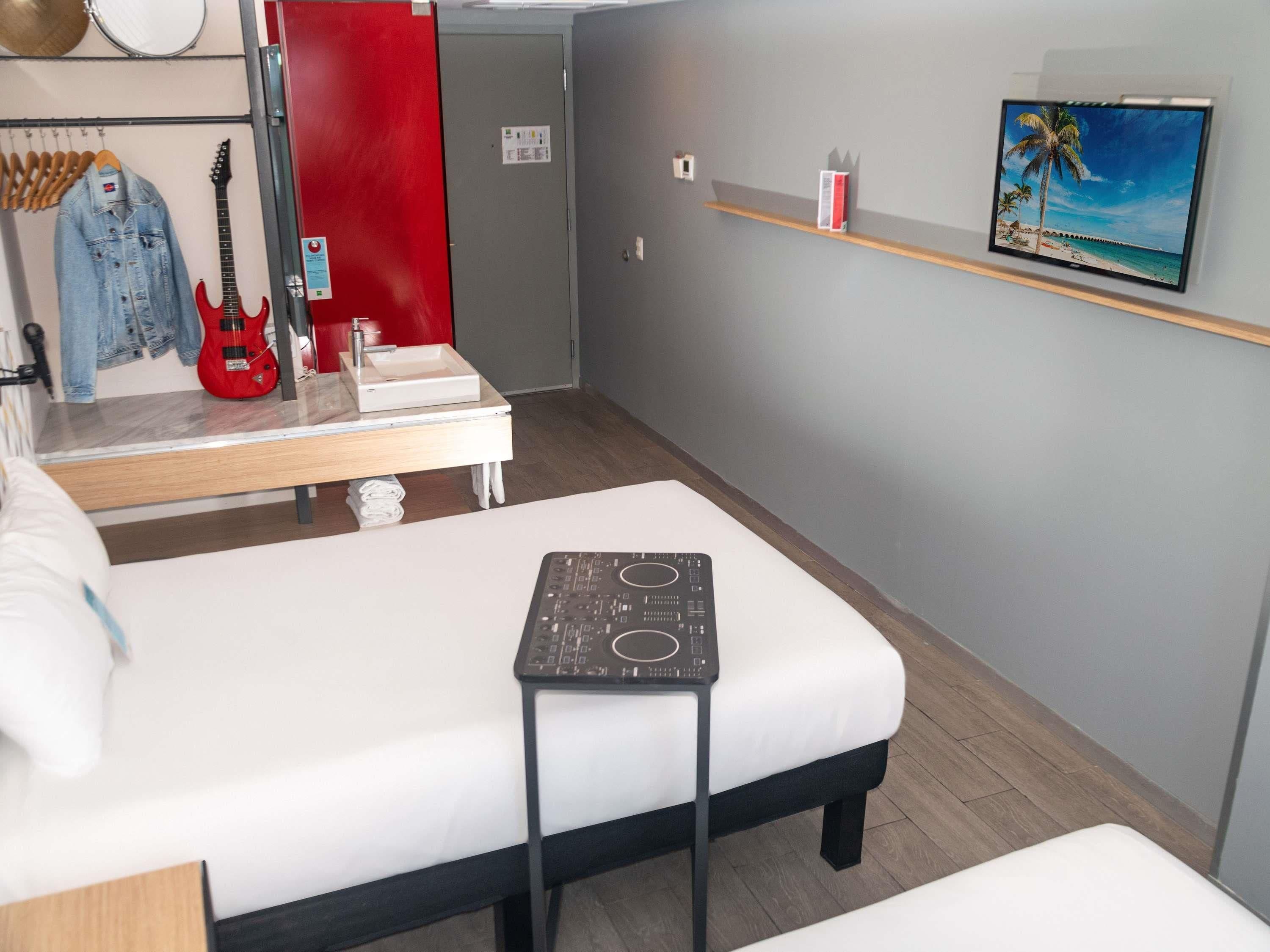 A hotel room with a bed, a small table, a red guitar, a mirror, a red door, a shelf with a picture, and a wall-mounted TV.