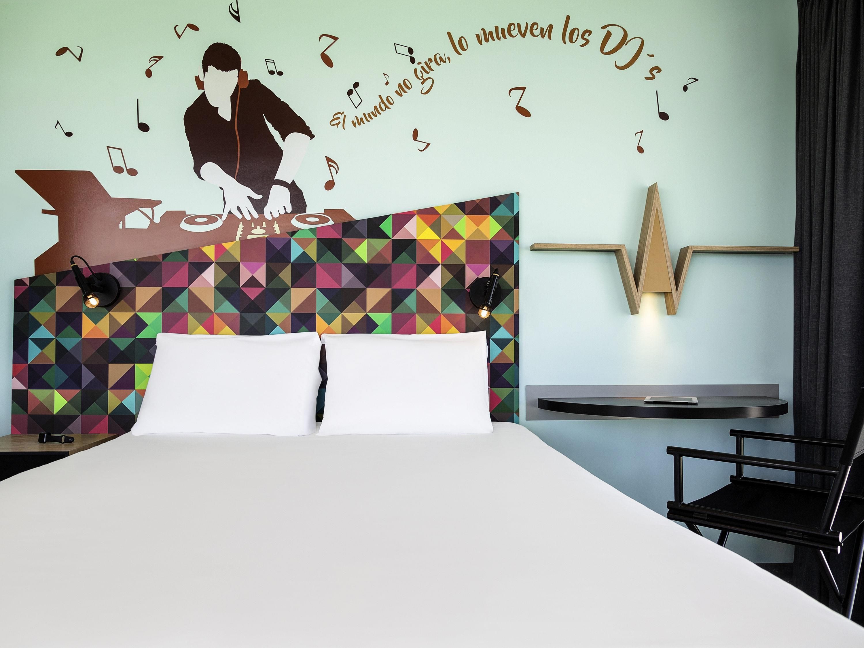 A hotel room with a bed, a desk, a chair, a wall with a geometric pattern, and a wall-mounted light fixture.