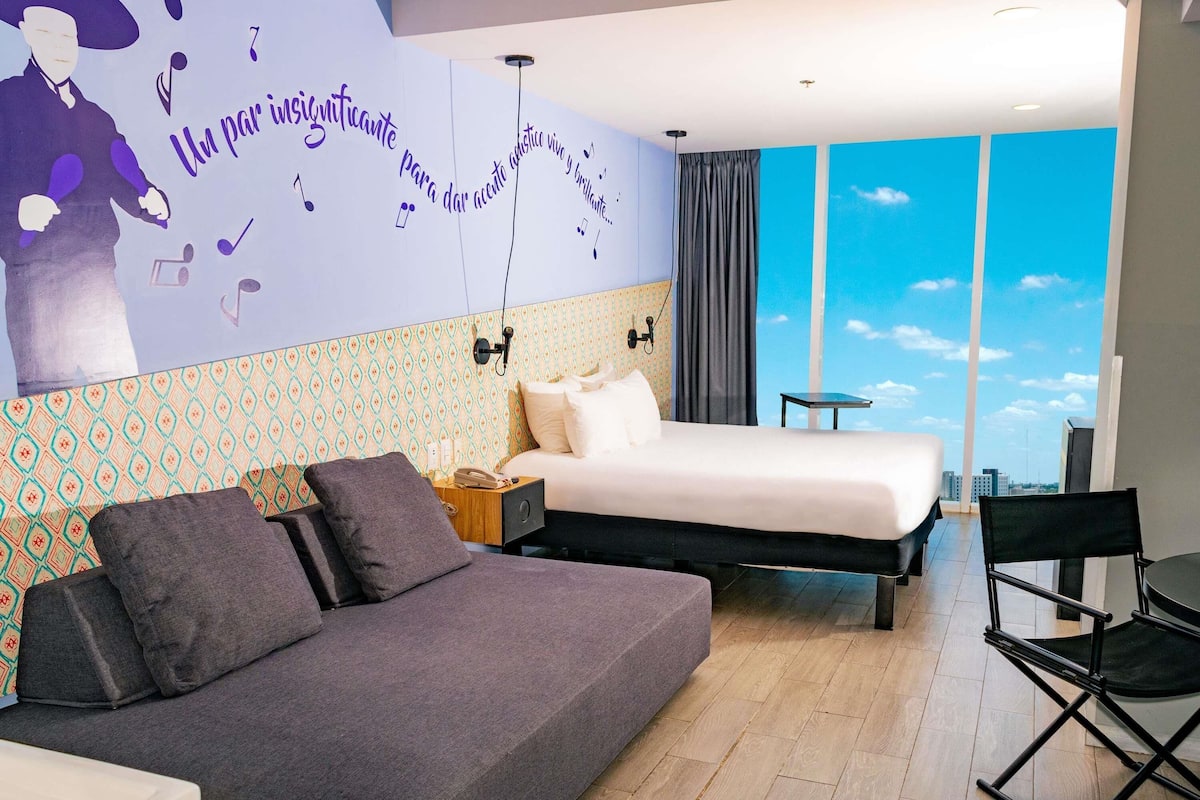 Ein modernes Hotelzimmer mit einem großen Bett, einer Sitzecke, einem Schreibtisch und Blick zum Himmel durch große Fenster.