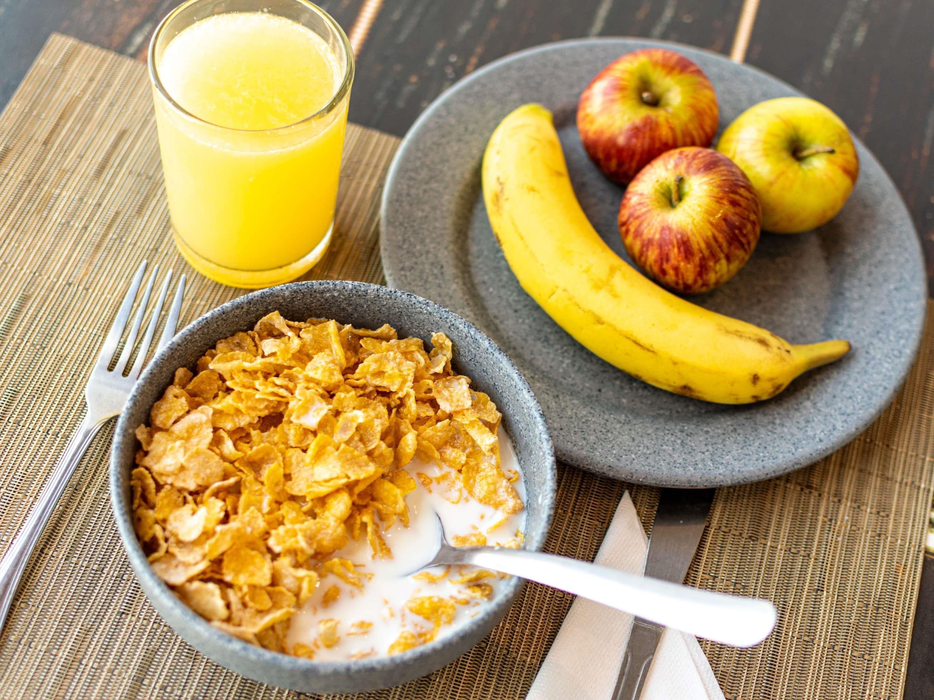 Un desayuno con cereal, un vaso de zumo de naranja y un plato de plátanos y manzanas.