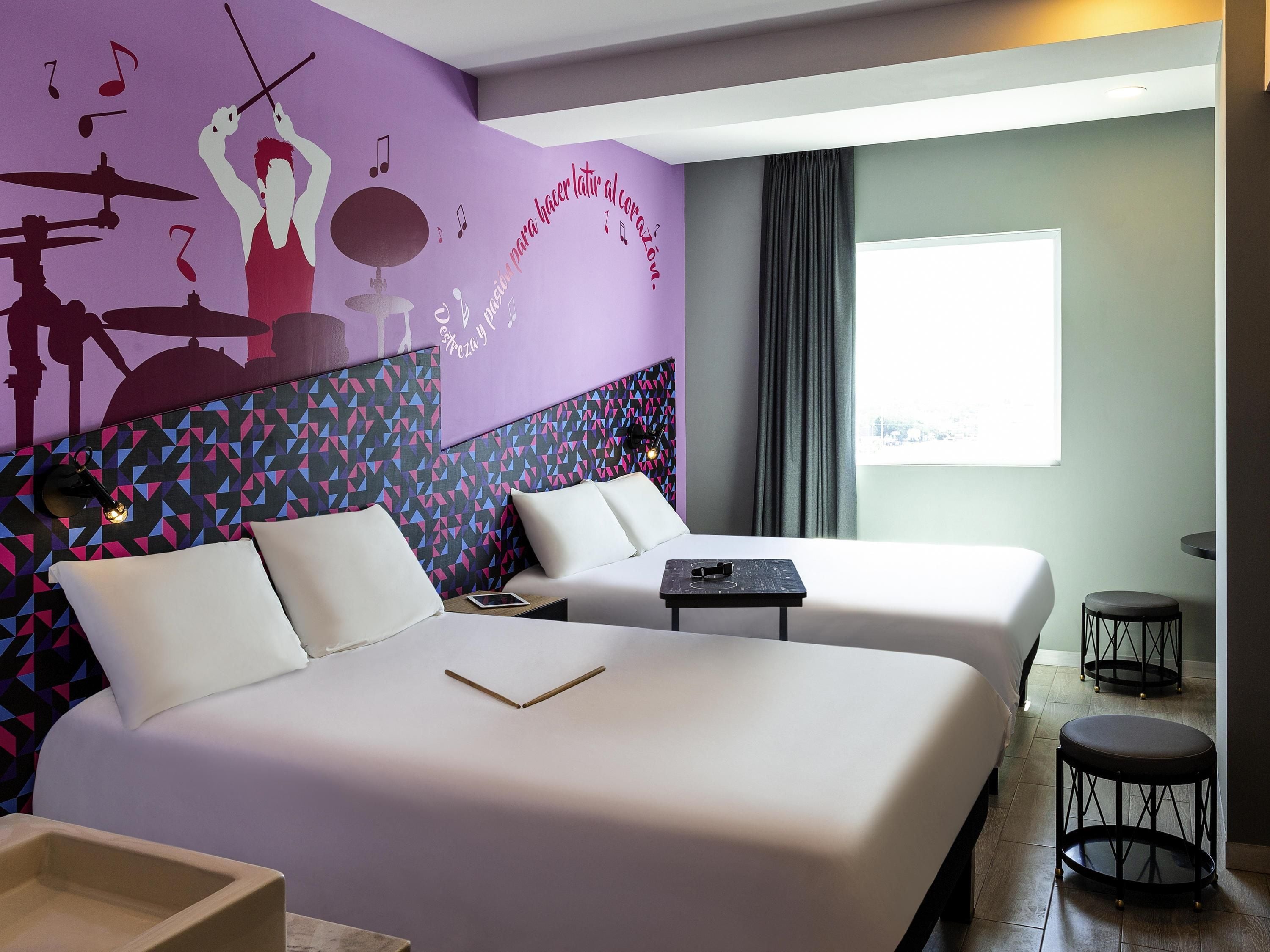 A hotel room with two beds, a wall mural, a window, and a small table.